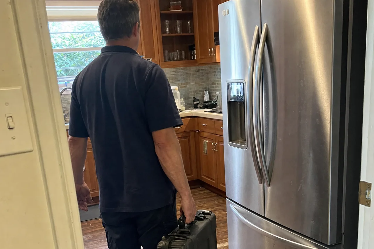 Technician carrying a tool case into a timber-cabinet kitchen, walking toward a stainless refrigerator