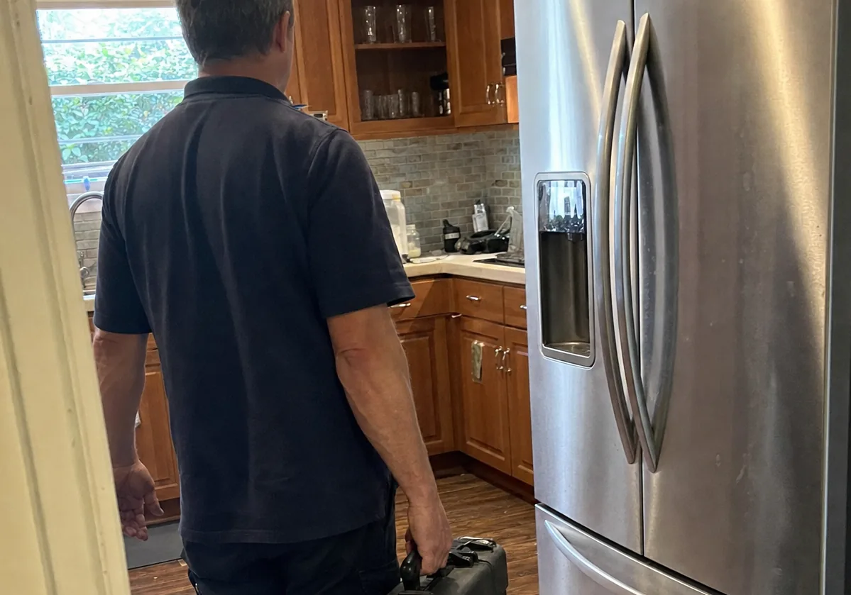 Centred frame of a technician entering a wood-panelled kitchen, a stainless refrigerator ahead