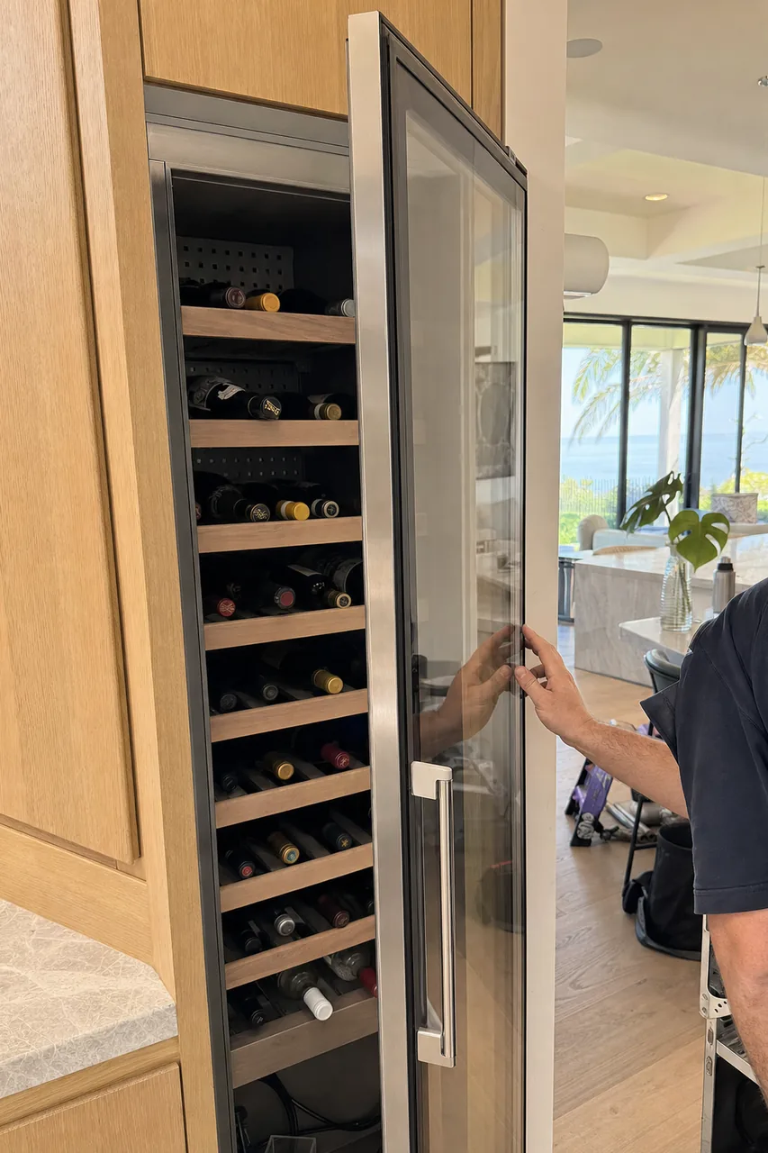 Glass-fronted wine column standing open on racked bottles, a hand steadying the door frame